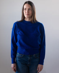 A woman with long brown hair wears the White & Warren Cashmere Crew in Frontier Blue with blue jeans, standing against a plain light-colored background and looking slightly to her left.