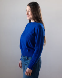 A woman with long brown hair, wearing the White & Warren Cashmere Crew in Frontier Blue and blue jeans, stands sideways and smiles against a plain light gray background.
