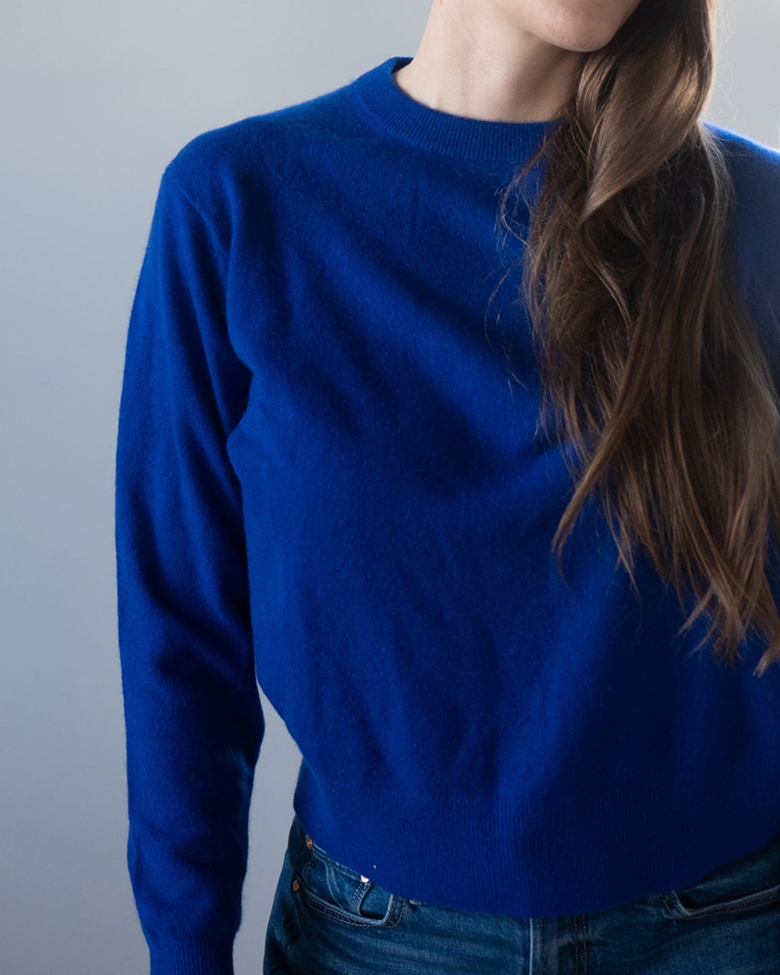 A person with long brown hair wears the White & Warren Cashmere Crew in Frontier Blue and blue jeans, standing against a plain light gray background. Only the upper body is visible, with the face partially out of frame.