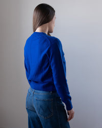 A woman with long brown hair wears the White & Warren Cashmere Crew in Frontier Blue and blue jeans, standing with her back and head turned slightly to the side against a plain, light-colored wall.