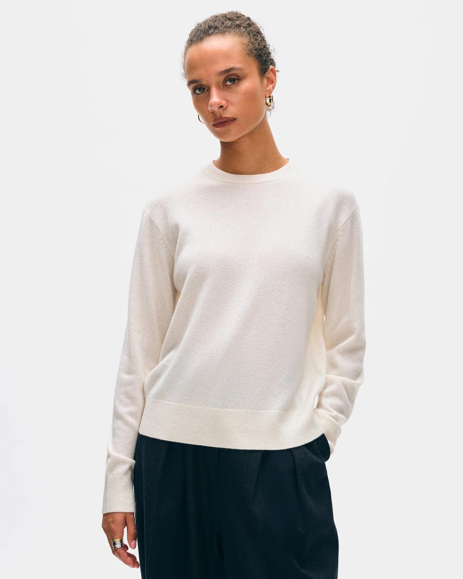 A person with curly hair in a bun wears the White & Warren Cashmere Crew in Soft White and dark pants, standing against a plain light background with a neutral expression.