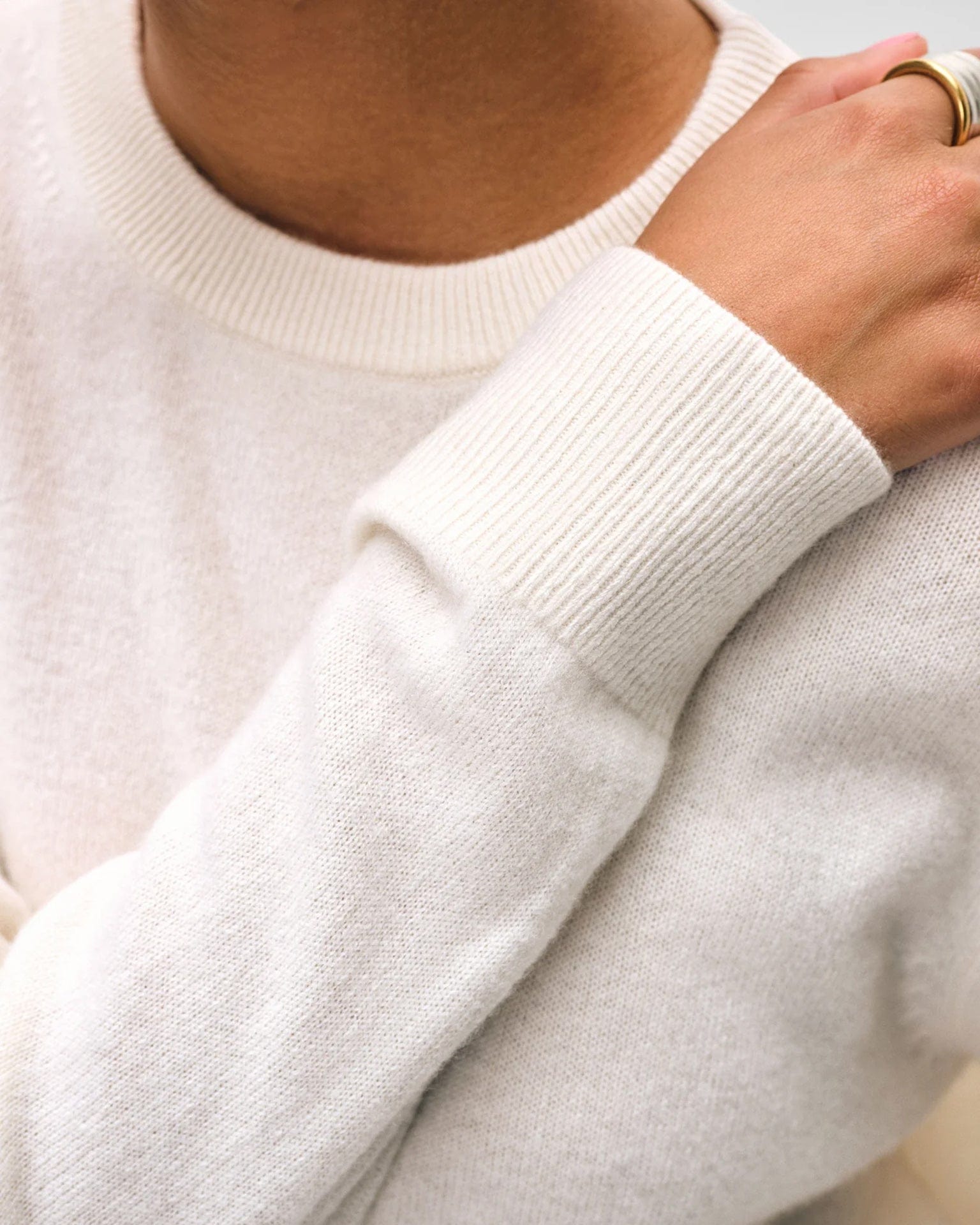 A person wearing the White & Warren Cashmere Crew in Soft White with ribbed cuffs rests their hand on their shoulder, showcasing gold rings.