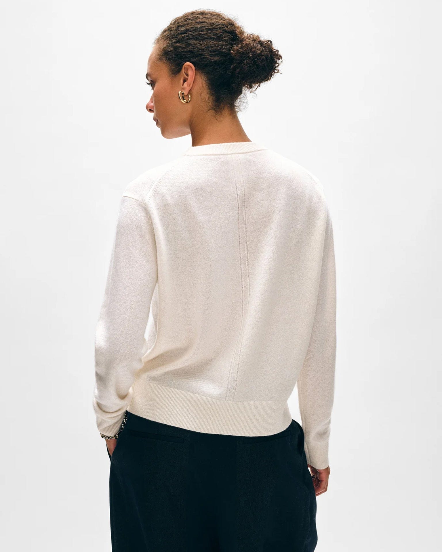 A person with curly hair in a bun is shown from the back, wearing the White & Warren Cashmere Crew in Soft White with dark pants, standing against a plain light background.