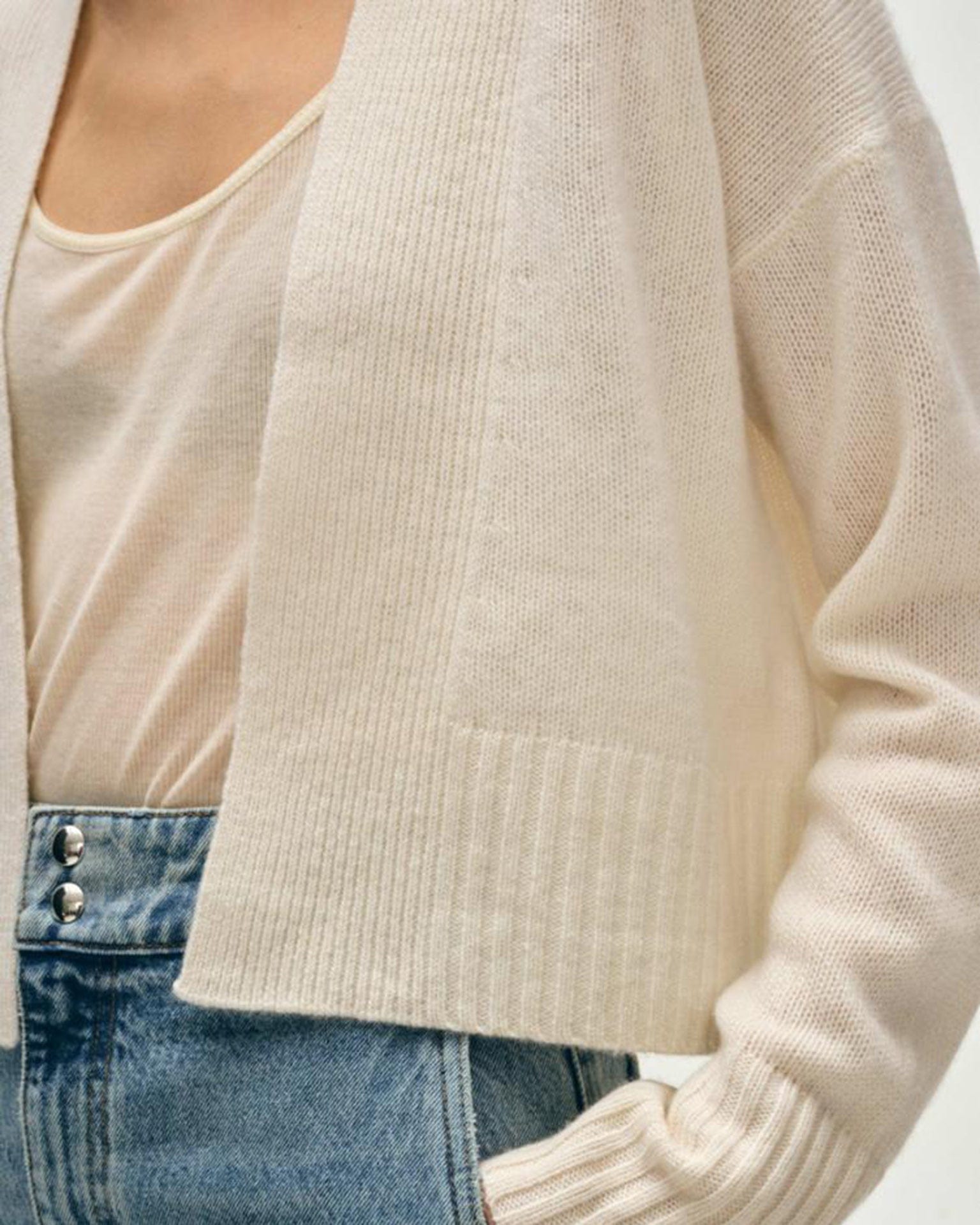 A person wears the White & Warren Cashmere Featherweight Open Cardigan in Soft White over a light beige top with high-waisted blue jeans. The image is cropped to show their torso and part of one arm.