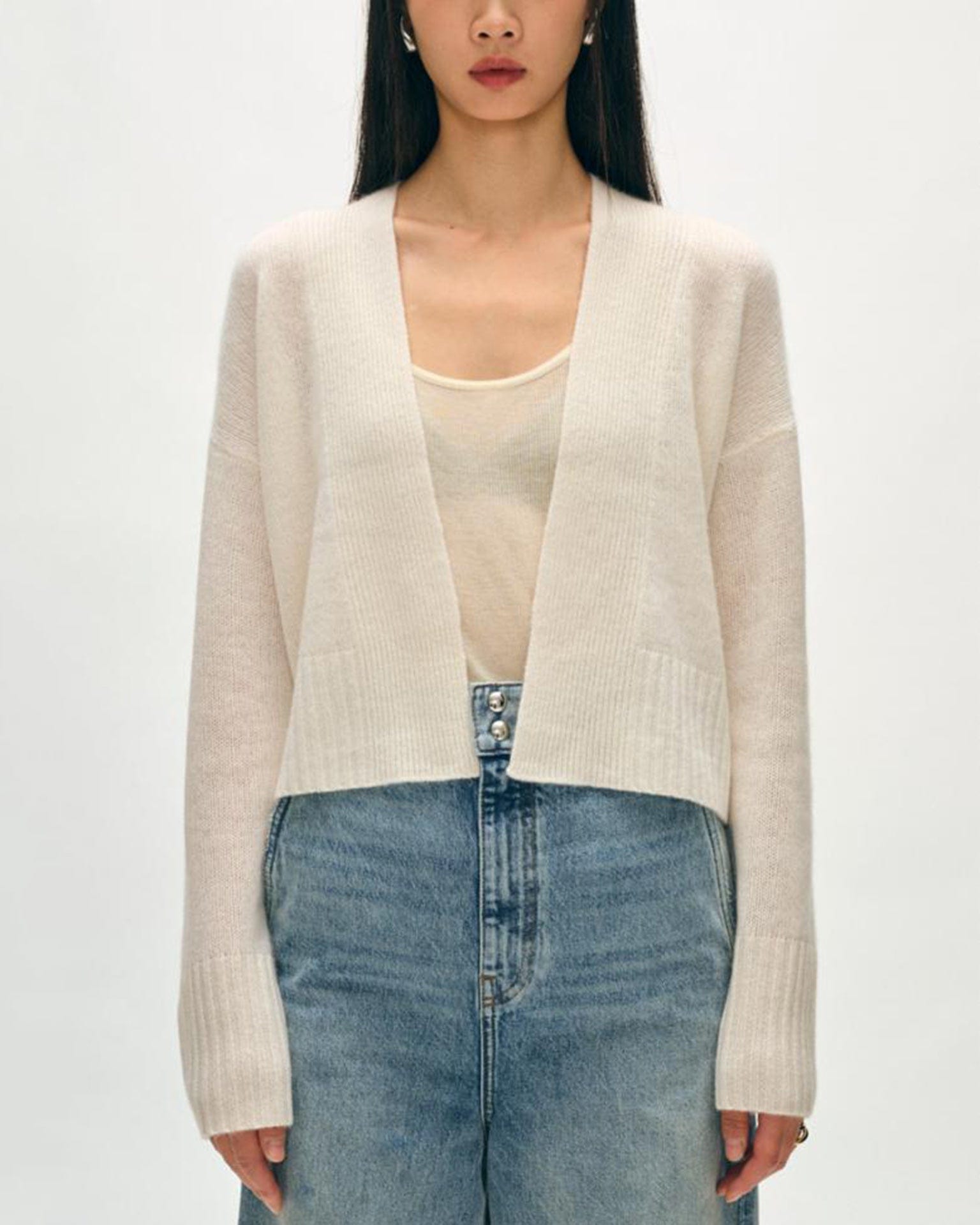 A woman wearing the White & Warren Cashmere Featherweight Open Cardigan in Soft White over a sheer beige tank top and high-waisted blue jeans stands against a plain light background.