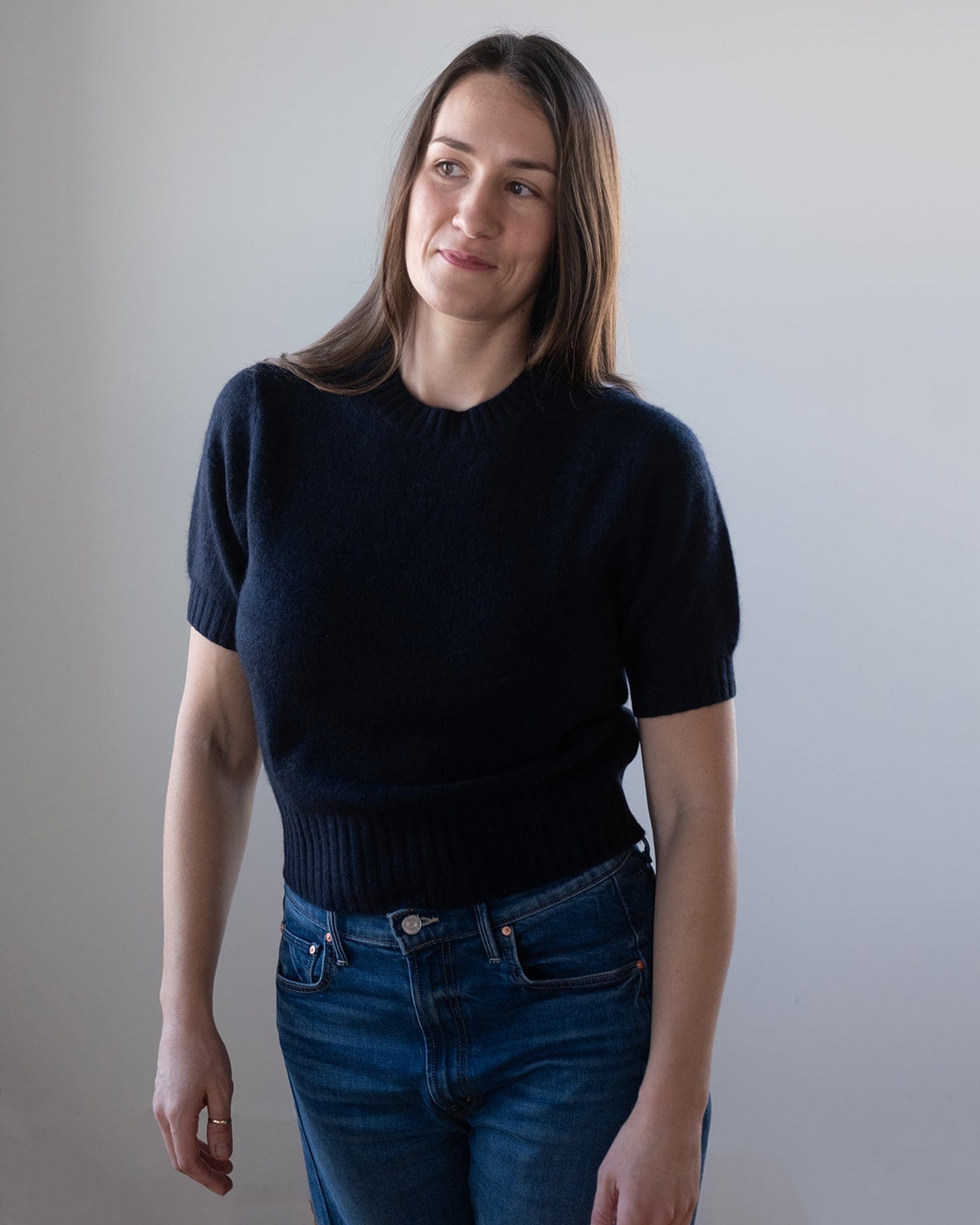 A woman with long brown hair models the White & Warren Cashmere Featherweight TShirt in Deep Navy with blue jeans, standing against a light background and looking slightly to the side with a soft smile.