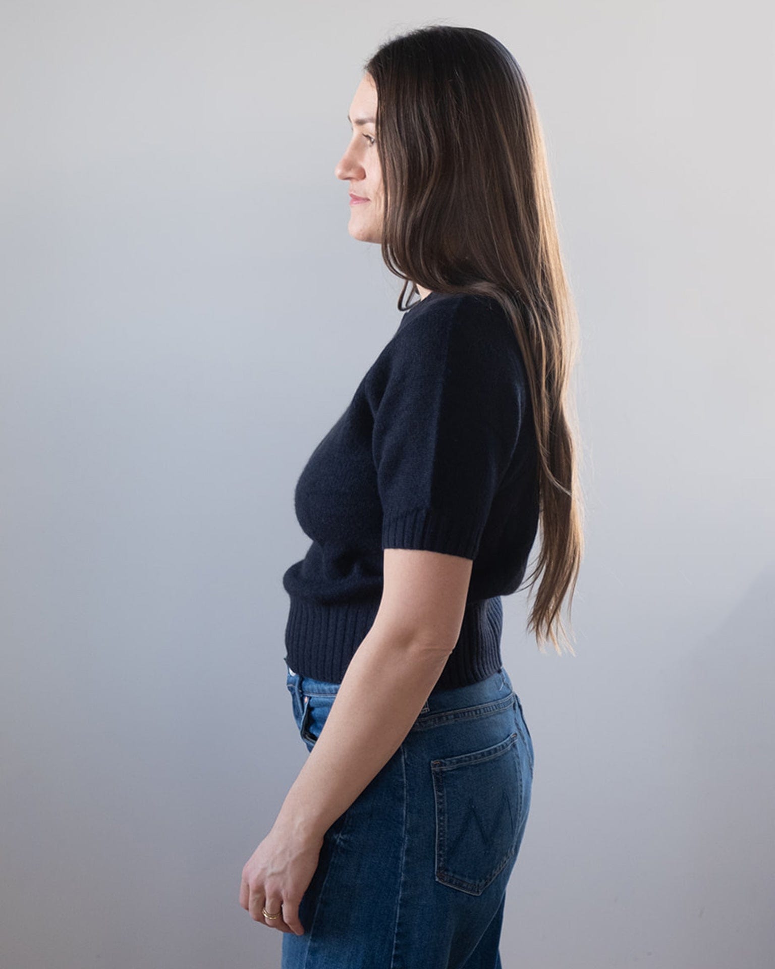 A woman with long brown hair, wearing a cropped Cashmere Featherweight TShirt in Deep Navy by White & Warren and blue jeans, stands in profile against a plain light background.