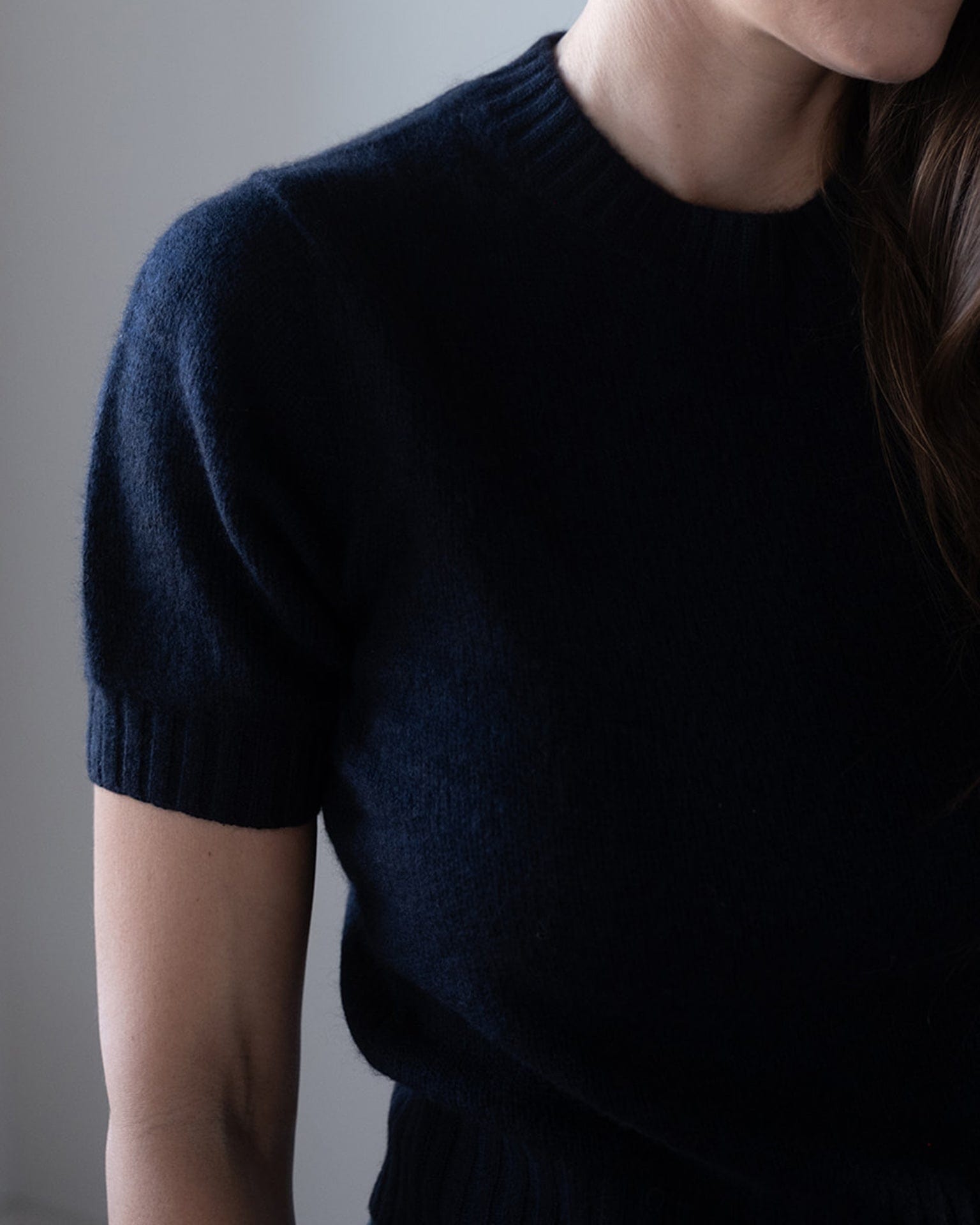 A person is shown from shoulders to waist, partially facing the camera and wearing the White & Warren Cashmere Featherweight TShirt in Deep Navy. Their face is mostly out of frame, and long brown hair appears on one side.