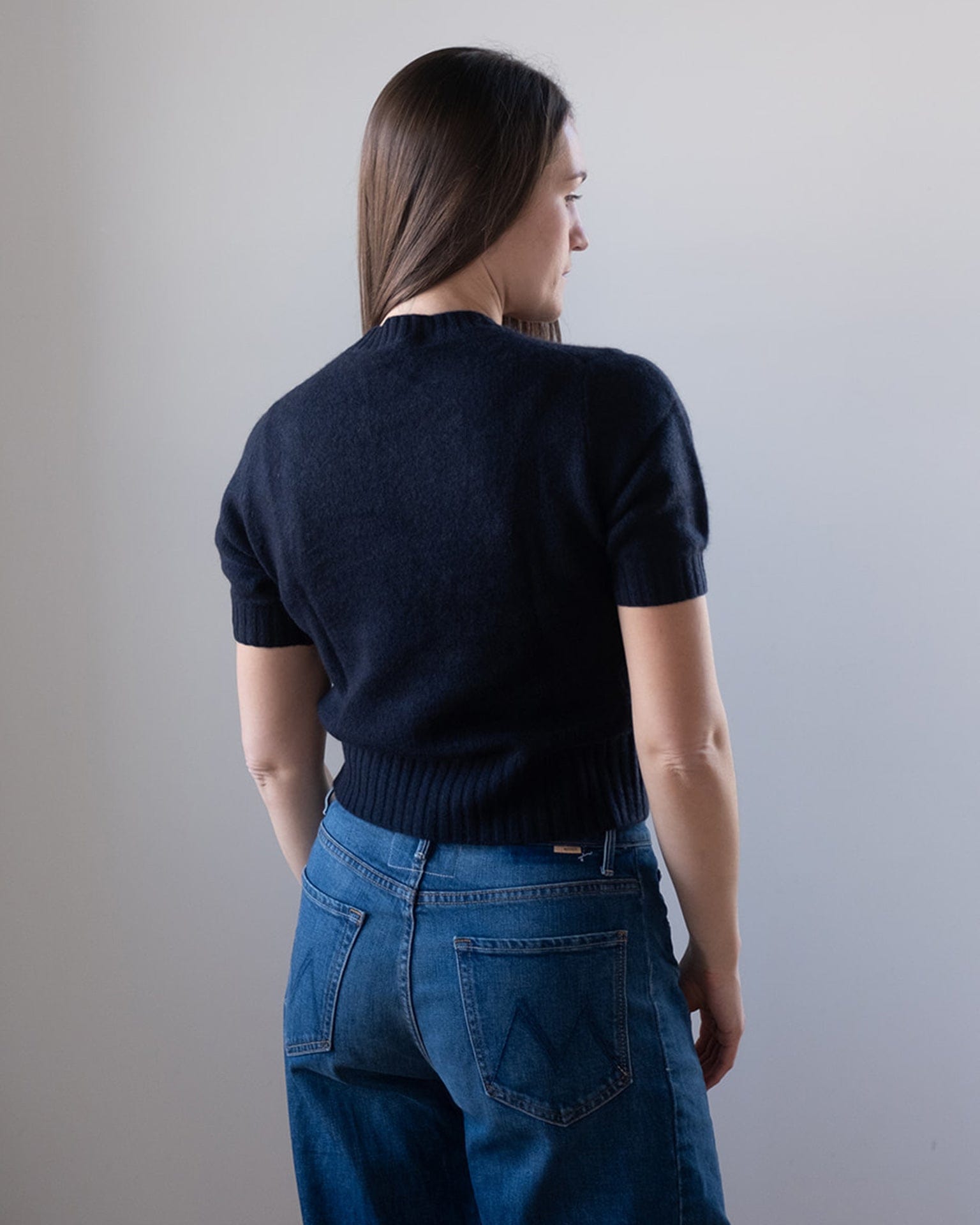 A woman with long brown hair faces away, wearing a White & Warren Cashmere Featherweight TShirt in Deep Navy and high-waisted blue jeans with large back pockets, set against a plain light-colored background.