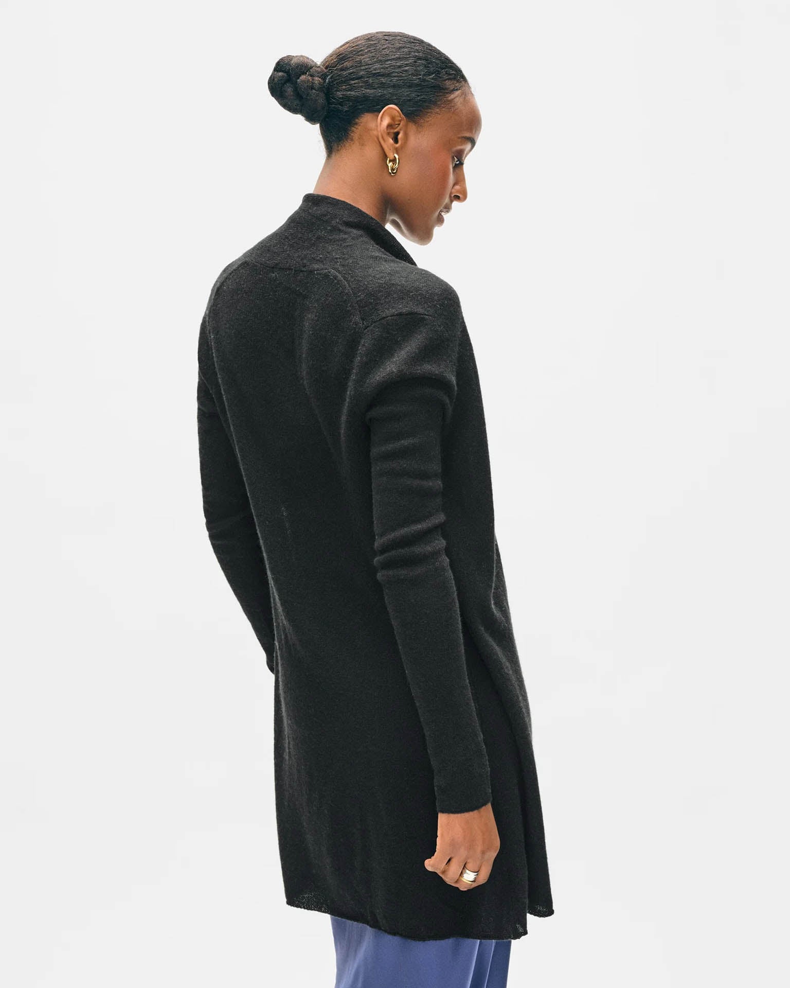 A woman with dark hair in a bun models the White & Warren Cashmere Trapeze Cardigan in Black with blue pants, standing sideways and looking slightly down against a plain white background.