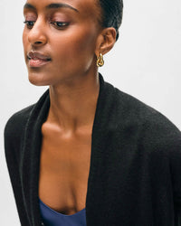 A woman with smooth brown skin, gold hoop earrings, and a blue top wears the White & Warren Cashmere Trapeze Cardigan in Black, looking slightly downward against a plain light background.