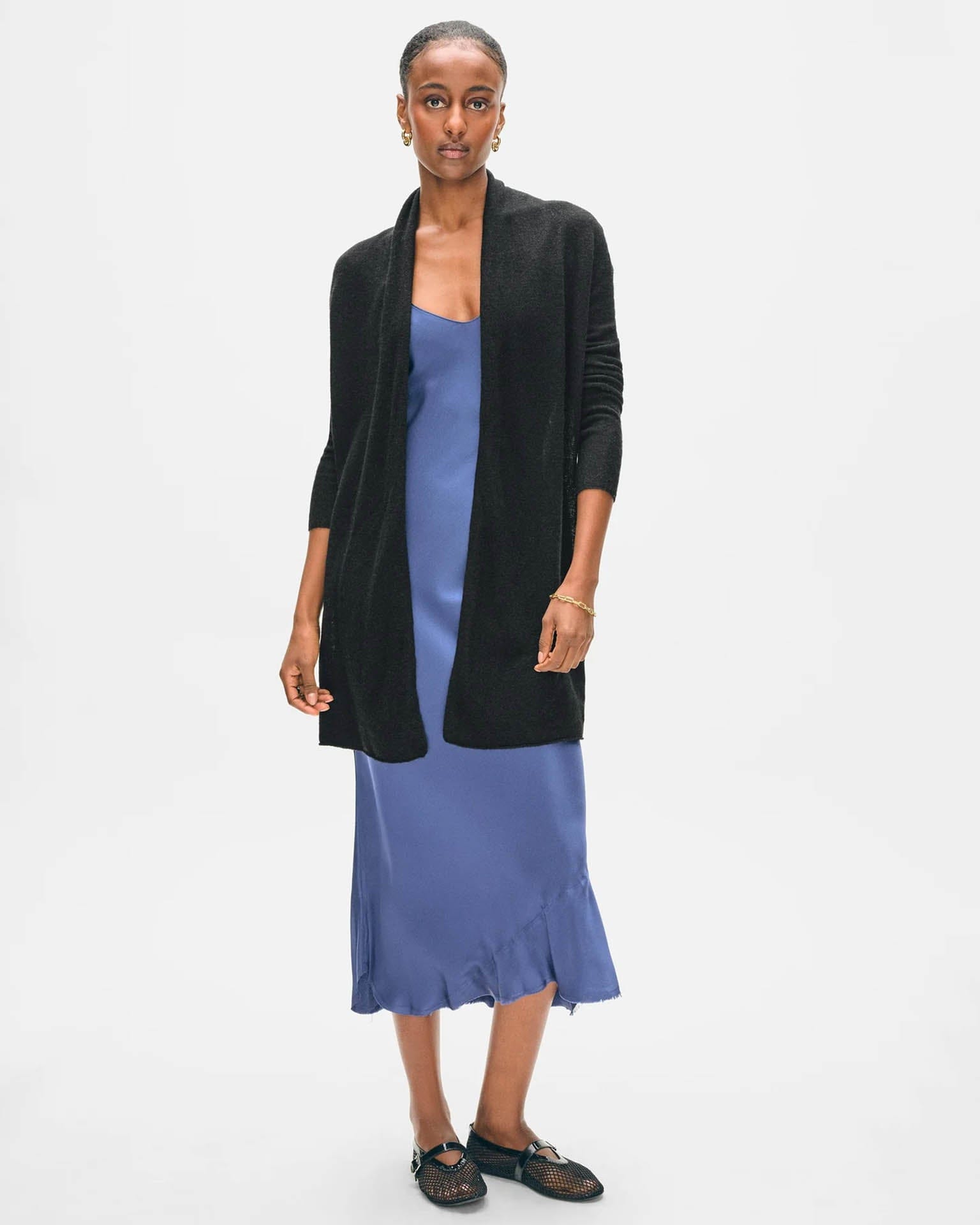 A woman wearing the White & Warren Cashmere Trapeze Cardigan in Black poses against a plain white background, styled with a blue satin midi dress, black woven flats, and simple jewelry, looking directly at the camera.