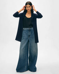 A woman with long dark hair stands facing forward, wearing a White & Warren Cashmere Trapeze Cardigan in Deep Navy over a black top and wide-leg blue jeans. Her hands are raised to her hair against a plain light gray background.