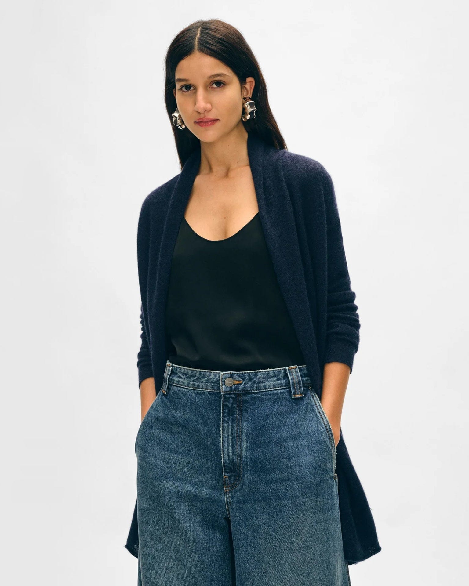 A woman with long dark hair wears a White & Warren Cashmere Trapeze Cardigan in Deep Navy over a black camisole and high-waisted blue jeans, standing with her hands in her pockets against a plain light background.