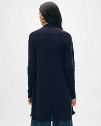 A person with long dark hair, wearing a White & Warren Cashmere Trapeze Cardigan in Deep Navy and blue jeans, stands facing away from the camera against a plain light background.