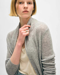 A woman with a short blonde bob wears the White & Warren Cashmere Trapeze Cardigan in Grey Heather over a cream top. She looks at the camera with a neutral expression, holding the collar and displaying rings, against a plain light background.