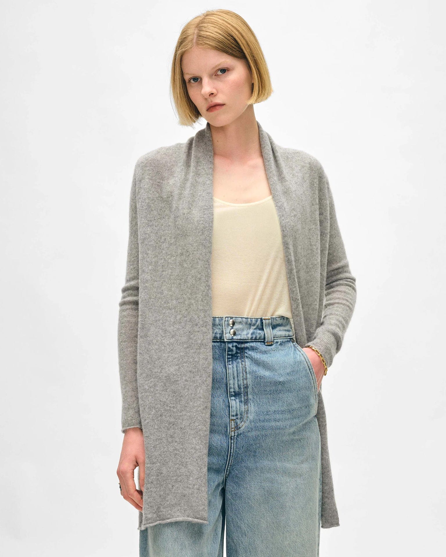 A woman with short blonde hair wears the White & Warren Cashmere Trapeze Cardigan in Grey Heather over a cream tank top and high-waisted blue jeans, standing with one hand in her pocket against a plain light background.