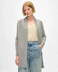 A woman with short blonde hair wears the White & Warren Cashmere Trapeze Cardigan in Grey Heather over a cream tank top and high-waisted blue jeans, standing with one hand in her pocket against a plain light background.