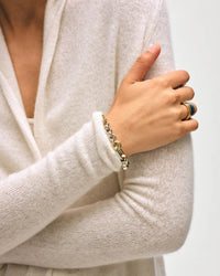 A person in the White & Warren Cashmere Trapeze Cardigan in Soft White crosses their arms, showing a gold and silver bracelet and two stacked rings—one black and gold—against a plain, light background.