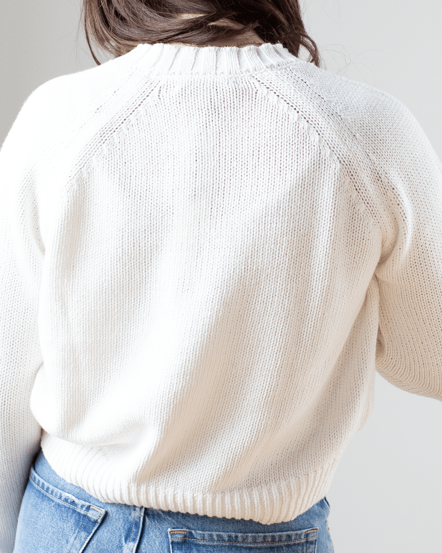 A person with long brown hair is seen from the back wearing the White & Warren Cotton Blend Beachy Crew in White and blue jeans. The well-lit scene highlights the relaxed fit and textured cotton linen blend of the sweater.