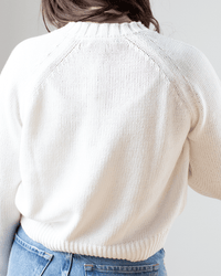 A person with long brown hair is seen from the back wearing the White & Warren Cotton Blend Beachy Crew in White and blue jeans. The well-lit scene highlights the relaxed fit and textured cotton linen blend of the sweater.