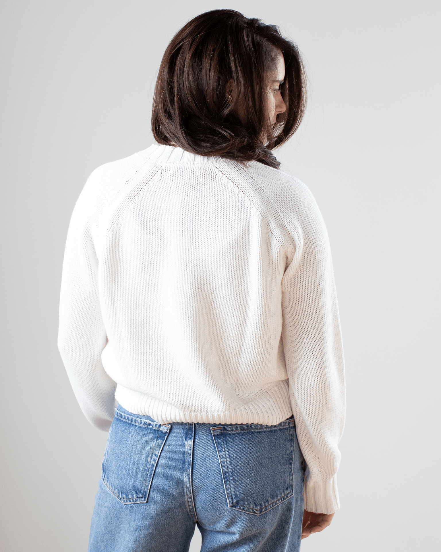 A person with shoulder-length brown hair, seen from behind, wears the White & Warren Cotton Blend Beachy Crew in white with blue jeans against a plain light gray background.