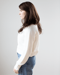 A woman with long brown hair, wearing a relaxed fit White & Warren Cotton Blend Beachy Crew in White and blue jeans, stands in profile against a plain light gray background.
