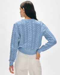 A woman with long dark hair stands facing away, wearing a White & Warren Cotton Slub Cable Crew in Soft Blue and high-waisted white pants against a plain light background.