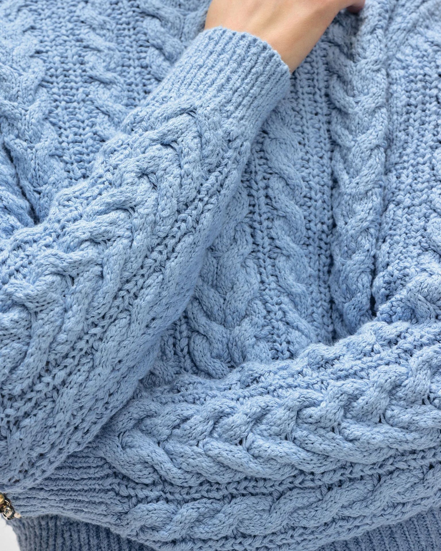 A close-up of a person wearing the White & Warren Cotton Slub Cable Crew in Soft Blue, with one arm crossed to highlight the sweater’s chunky cable knit texture.