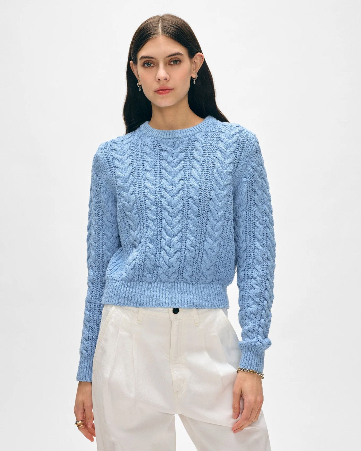 A woman with straight dark hair wears the White & Warren Cotton Slub Cable Crew in Soft Blue and high-waisted white pants against a plain light background. She accessorizes with small hoop earrings and a bracelet, her expression neutral.