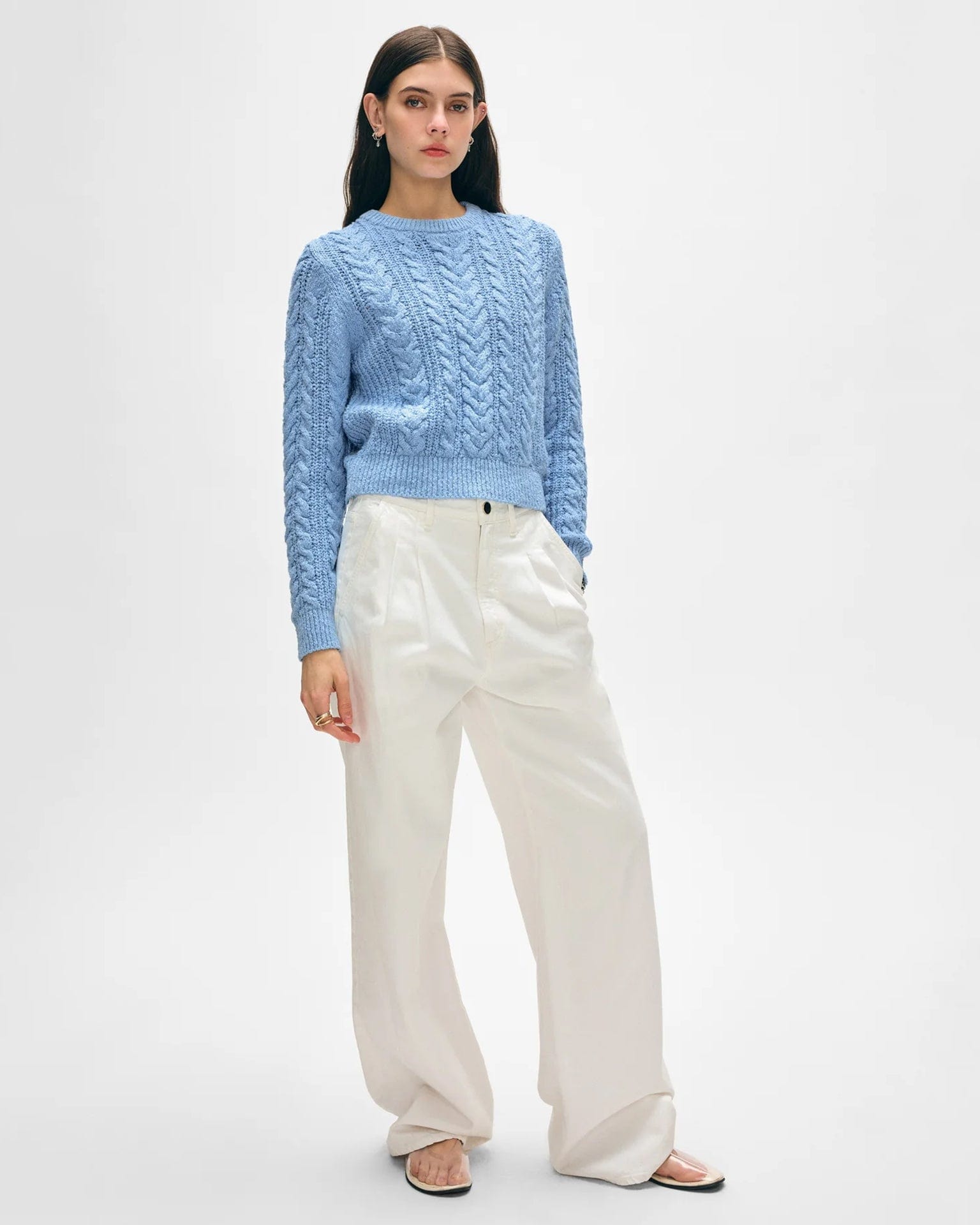 A woman in a White & Warren Cotton Slub Cable Crew in Soft Blue, loose white pants, and beige sandals stands against a plain background. She has long dark hair, a neutral expression, and one hand in her pocket.
