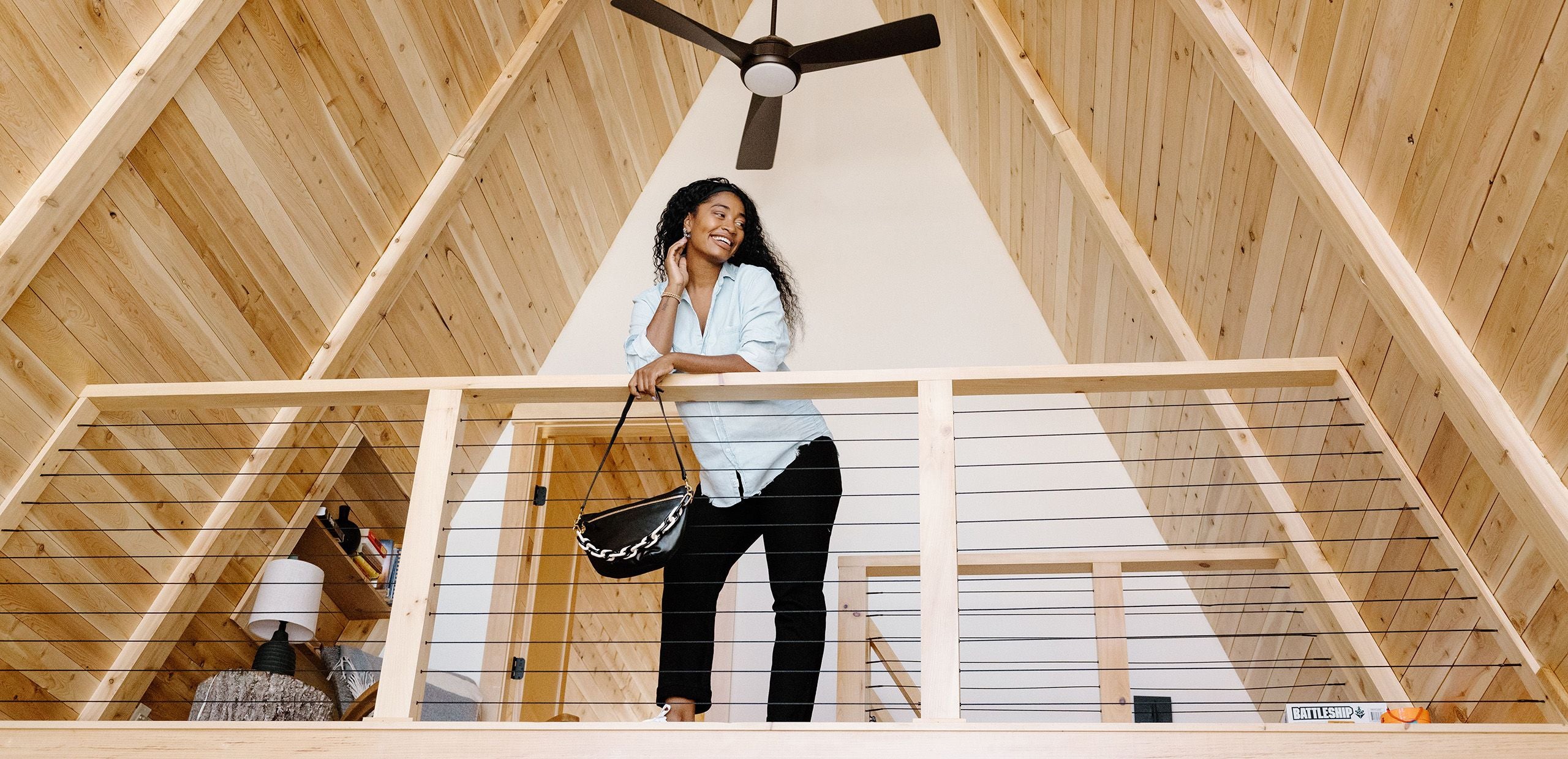 A woman leaning on the railing of a loft with a wooden ceiling and a fan above.