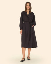 A woman stands against a beige background wearing the Caspy Dress in Black by XiRENA, a long-sleeved cotton poplin midi dress with ruffle collar details and a waist tie. She has straight, shoulder-length hair and black flats.