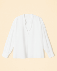 The XiRENA Emile Top in White is a collarless, long-sleeve cotton poplin blouse with a v-neck, shown laid flat on a light beige background.