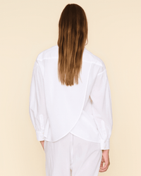 A person with long brown hair is seen from the back, wearing XiRENA’s Emile Top in White and white pants, standing against a beige background.