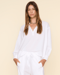 A woman with long brown hair wears a loose-fitting XiRENA Emile Top in White and white pants, standing against a light beige background with her hands in her pockets and a neutral expression.
