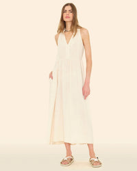A woman wears the XiRENA Liza Dress in Oysterette—a sleeveless, cream maxi with a V-neck and pockets—paired with beige sandals against a solid light background.