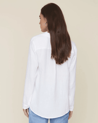 A woman with long brown hair, wearing the XiRENA Scout L/S Shirt in White and blue jeans, stands facing away from the camera against a plain beige background.