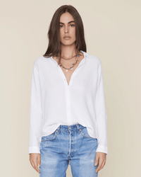 A person with long brown hair wears the XiRENA Scout L/S Shirt in White, partly tucked into light blue jeans and layered necklaces, standing against a plain beige background.