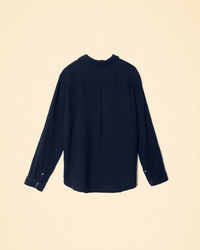 The XiRENA Scout Shirt in North Star, a women’s long sleeve shirt in navy cotton gauze, is shown flat on a beige background, highlighting its simple cuffs and subtly rounded back hem.