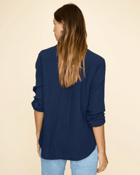 A woman with long brown hair, wearing the XiRENA Scout Shirt in North Star and light blue jeans, stands facing away from the camera against a beige background.