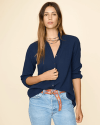 A woman with long brown hair wears a XiRENA Scout Shirt in North Star, light blue jeans, a brown patterned belt, and layered necklaces while standing against a plain beige background.