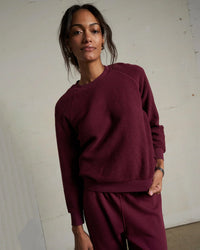 A woman in perfectwhitetee’s Ziggy Inside Out Fleece L/S Shrunken Crew SS in Dark Burgundy and matching pants stands indoors against a plain, light wall, looking confidently at the camera with a relaxed expression.