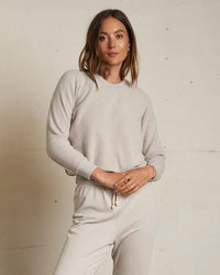 A woman confidently poses by a light wall wearing the perfectwhitetee Ziggy Inside Out Fleece L/S Shrunken Crew SS in Silver, her hands resting near her waist.
