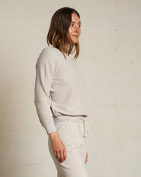 A woman with shoulder-length brown hair stands in profile against an off-white wall, wearing the perfectwhitetee Ziggy Inside Out Fleece L/S Shrunken Crew SS in Silver and matching drawstring pants, with a neutral expression.