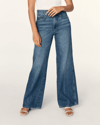 A person wearing the AMO Frida Wide Leg in Admire jeans, a white sleeveless top, and white heeled sandals stands against a plain light background. The person's head is not visible in the image.