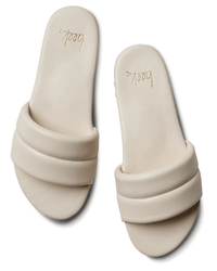 The Sugarbird in Eggshell by beek.. are cream-colored leather slide sandals with thick padded straps, gold beek.. branding on the insoles, and a cushioned memory foam footbed, shown against a white background.