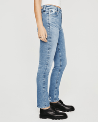 A person wearing AG Jeans' Alexxis Slim in 18Ys Presto, featuring a light blue wash and high-waisted, slim fit, paired with a white top and black chunky shoes, stands sideways against a plain white background.