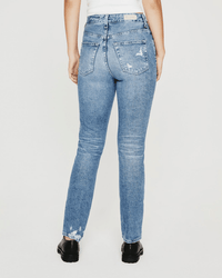 A person wearing AG Jeans' Alexxis Slim in 18Ys Presto—a high waisted, light blue distressed jean—black shoes, and a white top is seen from behind against a plain white background, emphasizing the jeans’ slim fit.