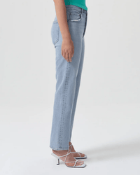 A person stands in profile against a white background wearing AGOLDE's High Rise Stovepipe in Destination jeans, a green top, and white strappy heeled sandals.